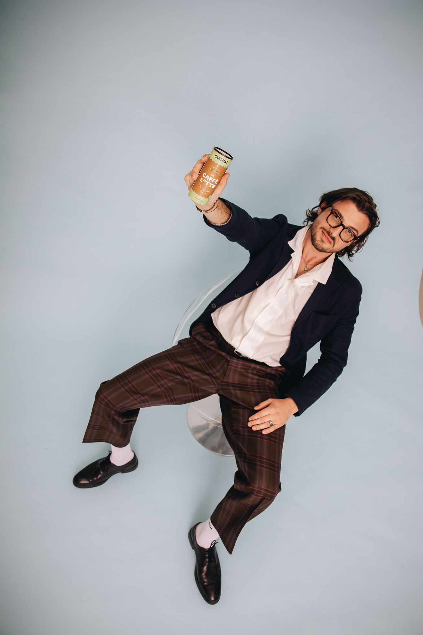 Ein junger Mann sitzt lässig auf einem transparenten Stuhl vor einem hellblauen Hintergrund. Er trägt eine Brille, ein weißes Hemd, eine dunkle Jacke und karierte Hosen. In seiner Hand hält er eine Hakuma Getränkedose, die er in die Kamera hält, als ob er sie präsentiert. Sein Ausdruck ist entspannt und selbstbewusst, während sein Outfit und die Pose einen modernen, stilvollen Eindruck vermitteln. Die Szene wirkt durch die Farbwahl und den minimalistischen Hintergrund modisch und fokussiert auf das Produkt in seiner Hand.